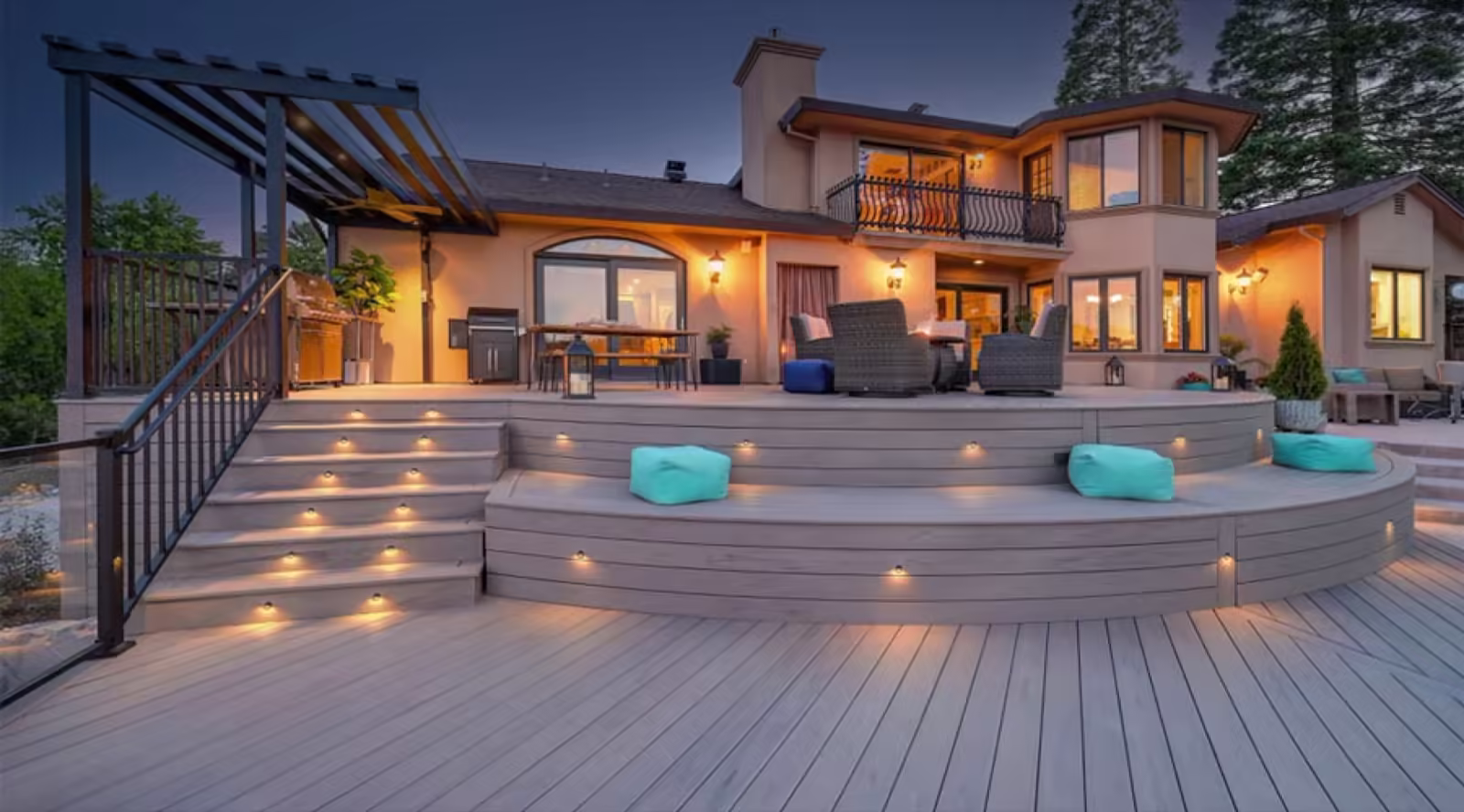 Curved grey composite deck with lit stair risers and tiered bench seating behind a two-storey home at dusk