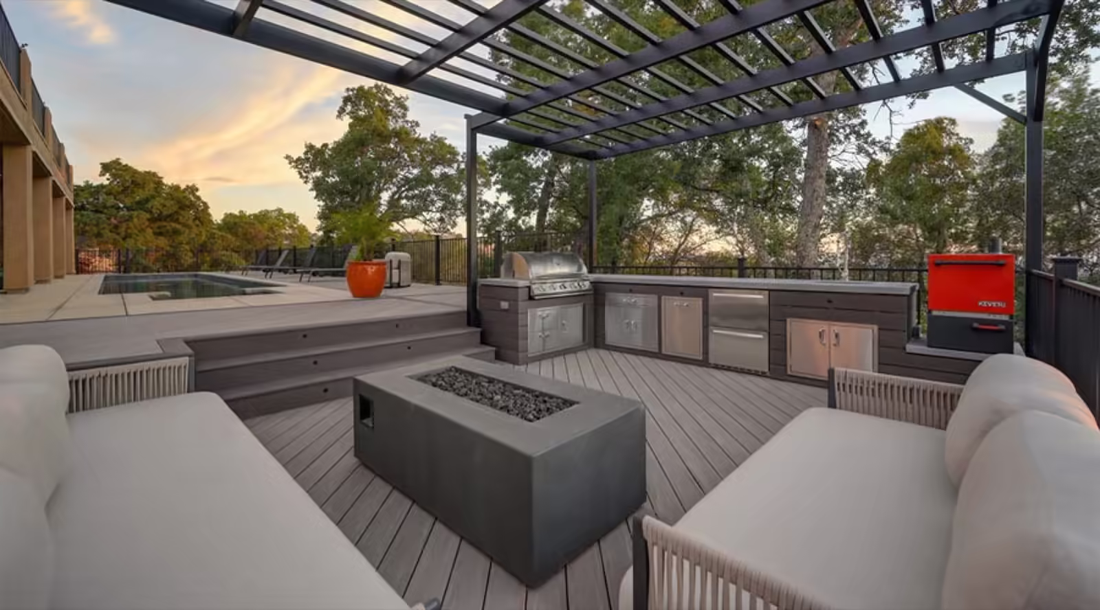 Composite deck under a steel pergola with an outdoor kitchen, gas fire table, lounge seating and a pool at sunset