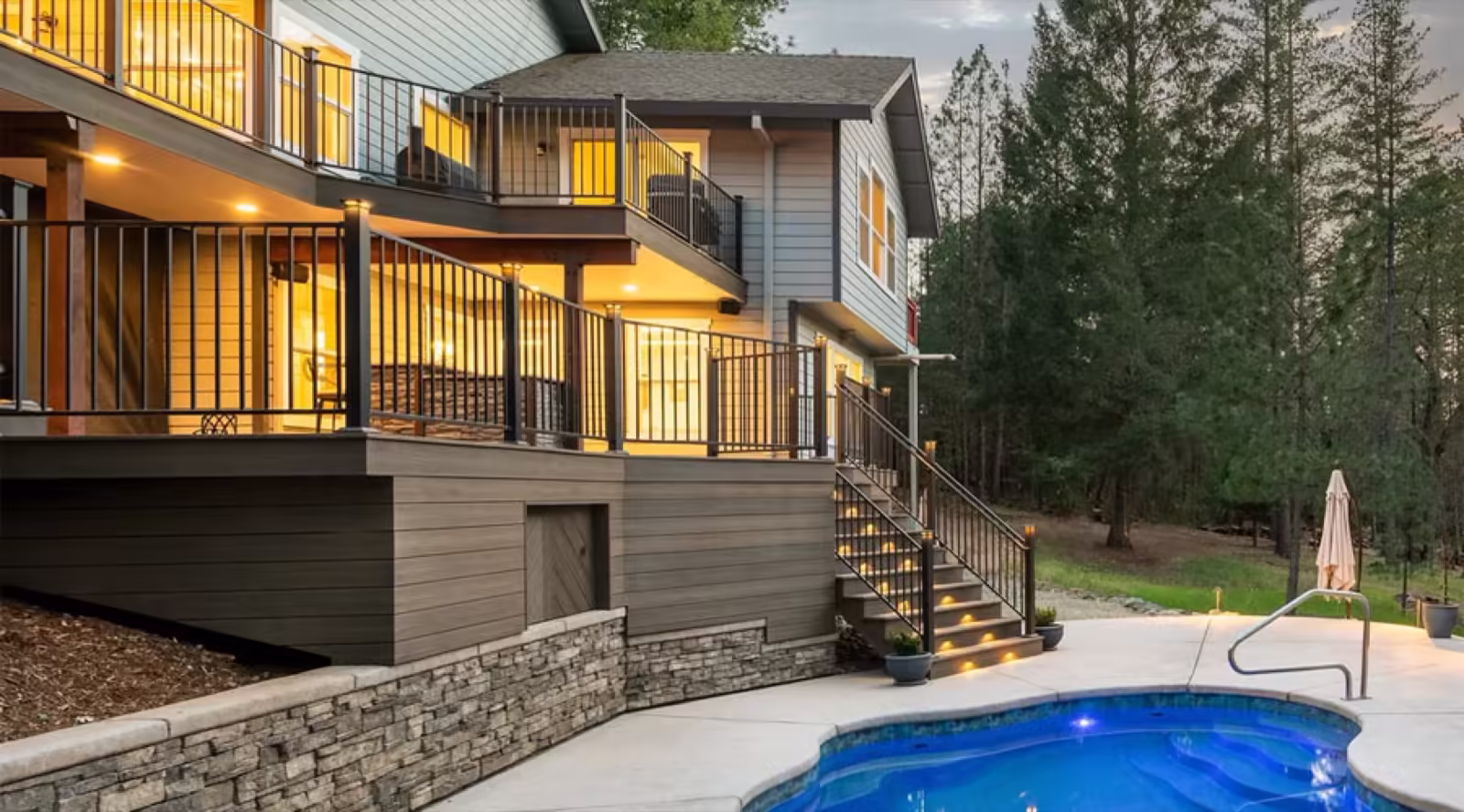 Two-storey deck and balcony with black metal railing and lit stairs running down to a pool and stone retaining wall