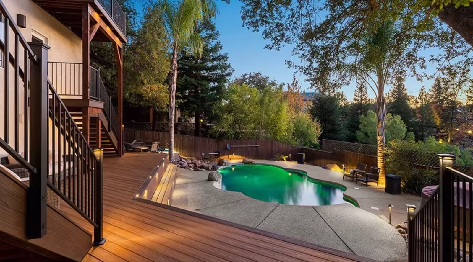 Two-level warm-toned deck with lit steps and black railing beside a lagoon-style swimming pool at dusk