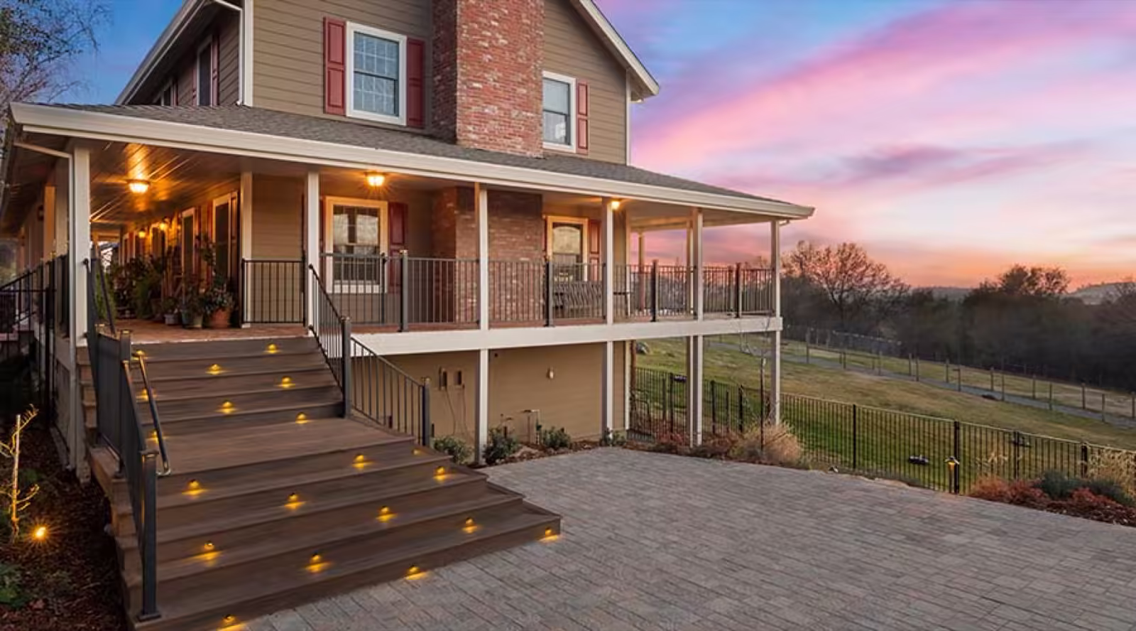 Covered wrap-around porch on a farmhouse with lit composite steps leading down to a paver patio at sunset