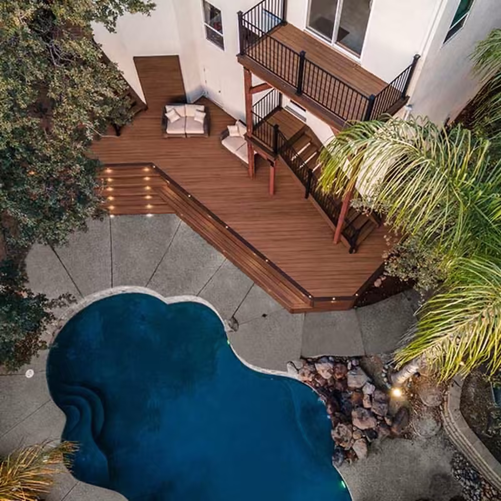 Overhead view of a brown composite deck wrapping a swimming pool, with a second-story balcony and black railings