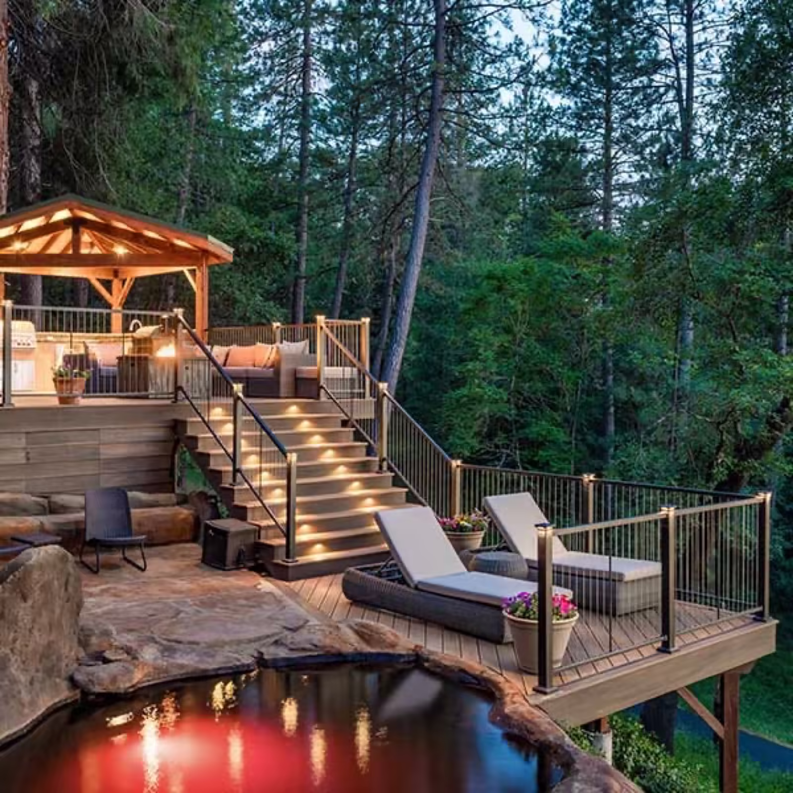 Multi-level deck at dusk in the pines with a lit gazebo, illuminated stairs and loungers beside a stone-edged pond